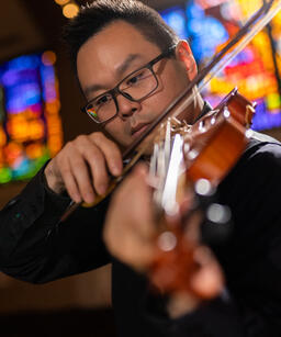 a man with glasses, black hair, and black shirt, playing the violin in front of a stained glass window